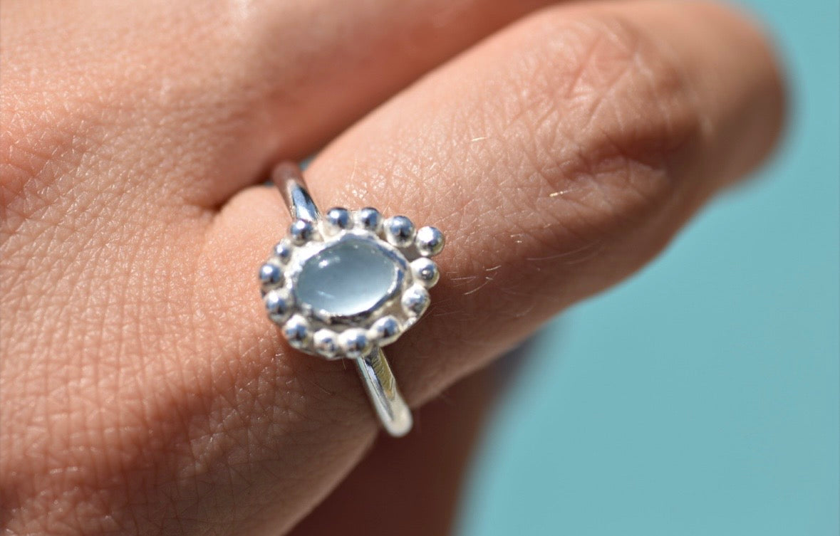 A sterling silver ring with a teardrop-shaped light blue gemstone surrounded by a beaded halo, worn on a finger against a solid light blue background.