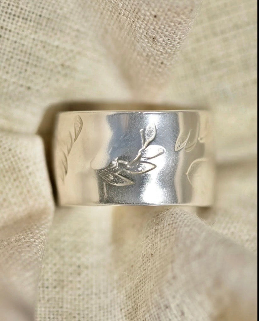 A wide silver band ring with engraved leaf motifs, displayed on a textured beige linen fabric background.