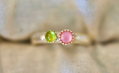 A delicate silver ring featuring two round cabochon gemstones, one green and one pink, displayed on a soft beige fabric background.