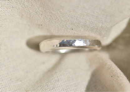 A hammered silver band ring displayed on a textured beige fabric stand.
